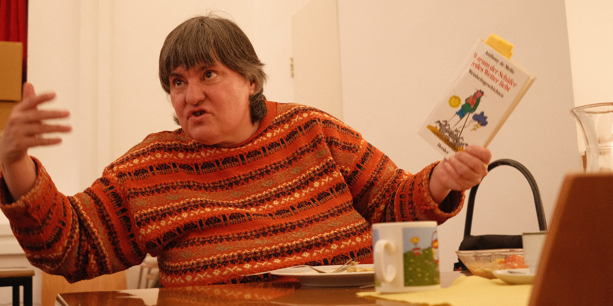 Eine Frau im orangen Pullover gestikuliert mit einem Buch in der Hand